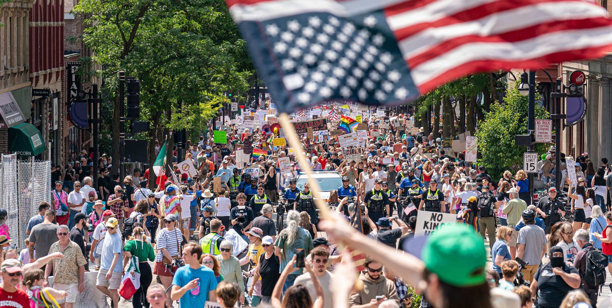 A crowded outdoor street protest or march with a large American flag in the foreground and many people walking down the road. Trees, buildings, and signs are visible throughout the scene.